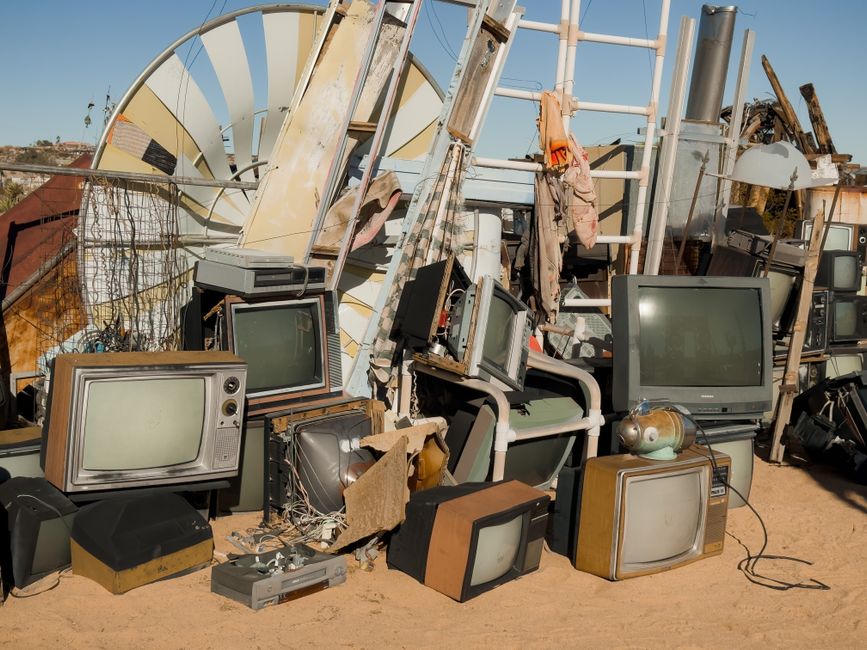 A large collection of discarded electronic devices, including multiple vintage television sets with wooden and plastic casings, all in various states of disrepair, piled together outdoors on a sandy s