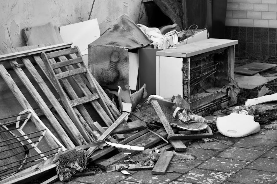 A large collection of discarded electronic devices, including multiple vintage television sets with wooden and plastic casings, all in various states of disrepair, piled together outdoors on a sandy s