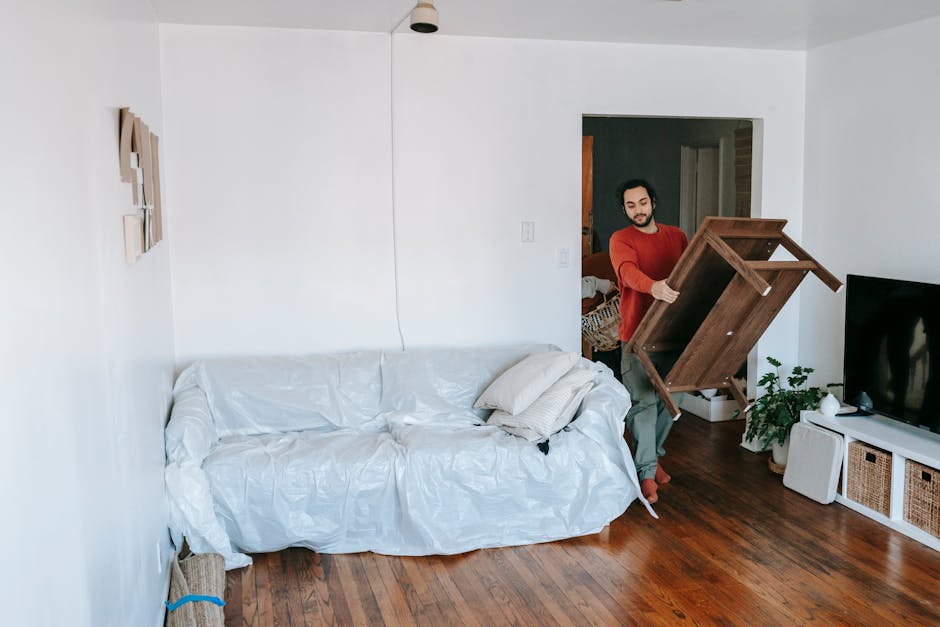 Two individuals are engaged in lifting and moving a large white sofa covered in protective wrapping, located in a bright, minimally decorated living room with white walls. The man, on the left, has da