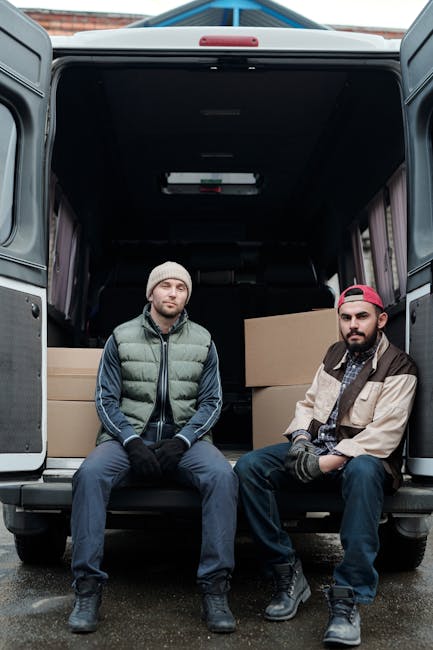 Two men sit in the open rear of a large black van parked outdoors on a paved surface, with visible brick buildings in the background. The man on the left wears a beige knit beanie, a puffy green and n
