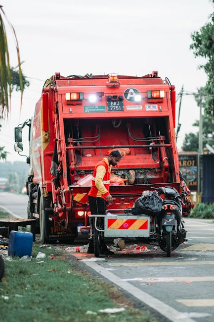 A sanitation worker wearing a yellow high-visibility vest and dark trousers is positioned behind a red rubbish collection vehicle on the side of a street. The worker appears to be inspecting or handli