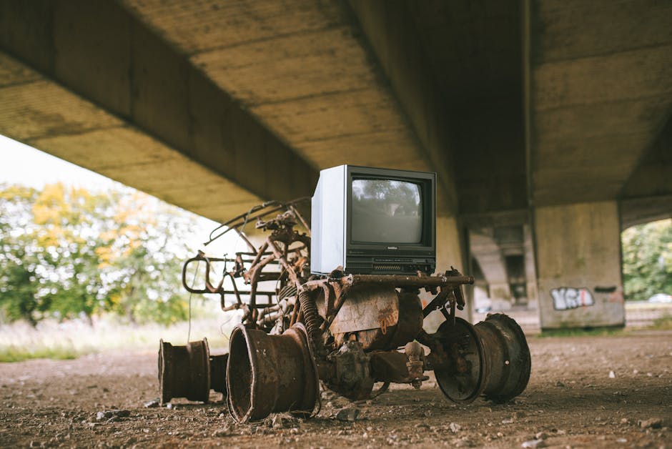 A discarded, broken microwave oven lying on the ground outdoors, surrounded by scattered debris, dirt, and patches of melting snow. The microwave's metallic and plastic exterior shows signs of dirt an