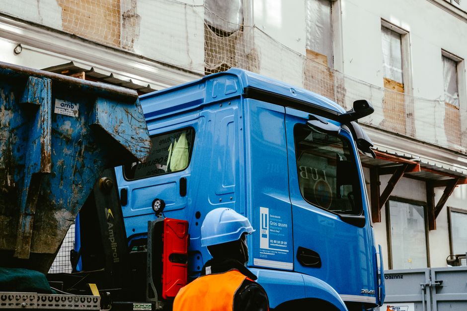 A bright blue rubbish collection truck with a large, metal hopper at the rear, situated on a city street. The truck's cabin features a black side mirror and a small, black door handle, with clear wind