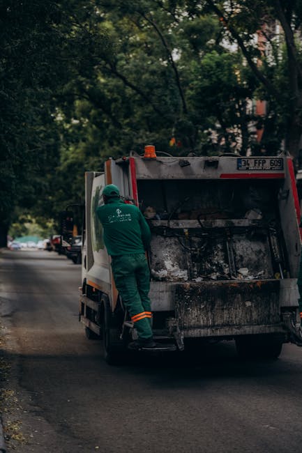 A bright blue rubbish collection truck with a large, metal hopper at the rear, situated on a city street. The truck's cabin features a black side mirror and a small, black door handle, with clear wind